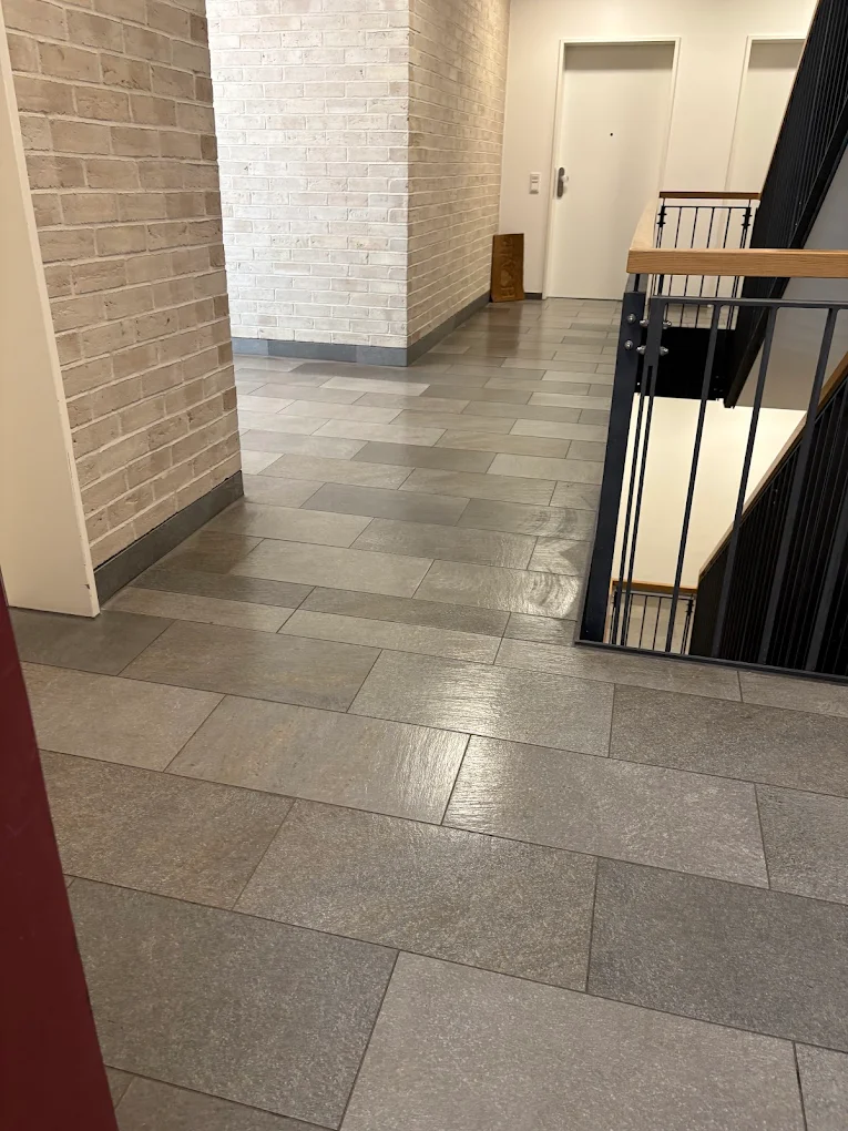 Innenansicht eines frisch gereinigten Flurs in einem Treppenhaus mit grauem Fliesenboden und Ziegelwand.