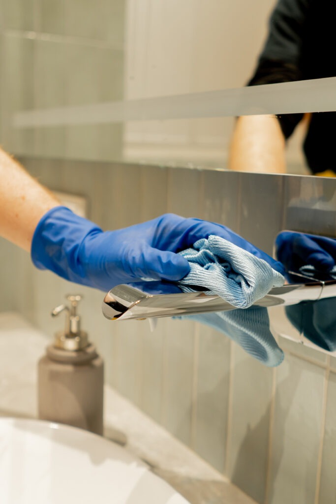Eine Hand in einem blauen Handschuh reinigt einen Chromhahn mit einem Tuch; Badezimmerreinigung in Hannover