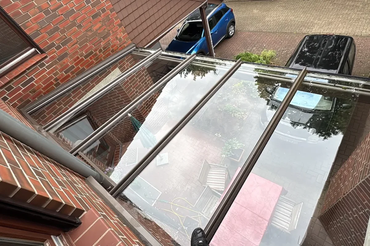 Blick von oben auf ein frisch gereinigtes Glasdach mit Metallrahmen, das die Terrasse und ein Auto spiegelt.