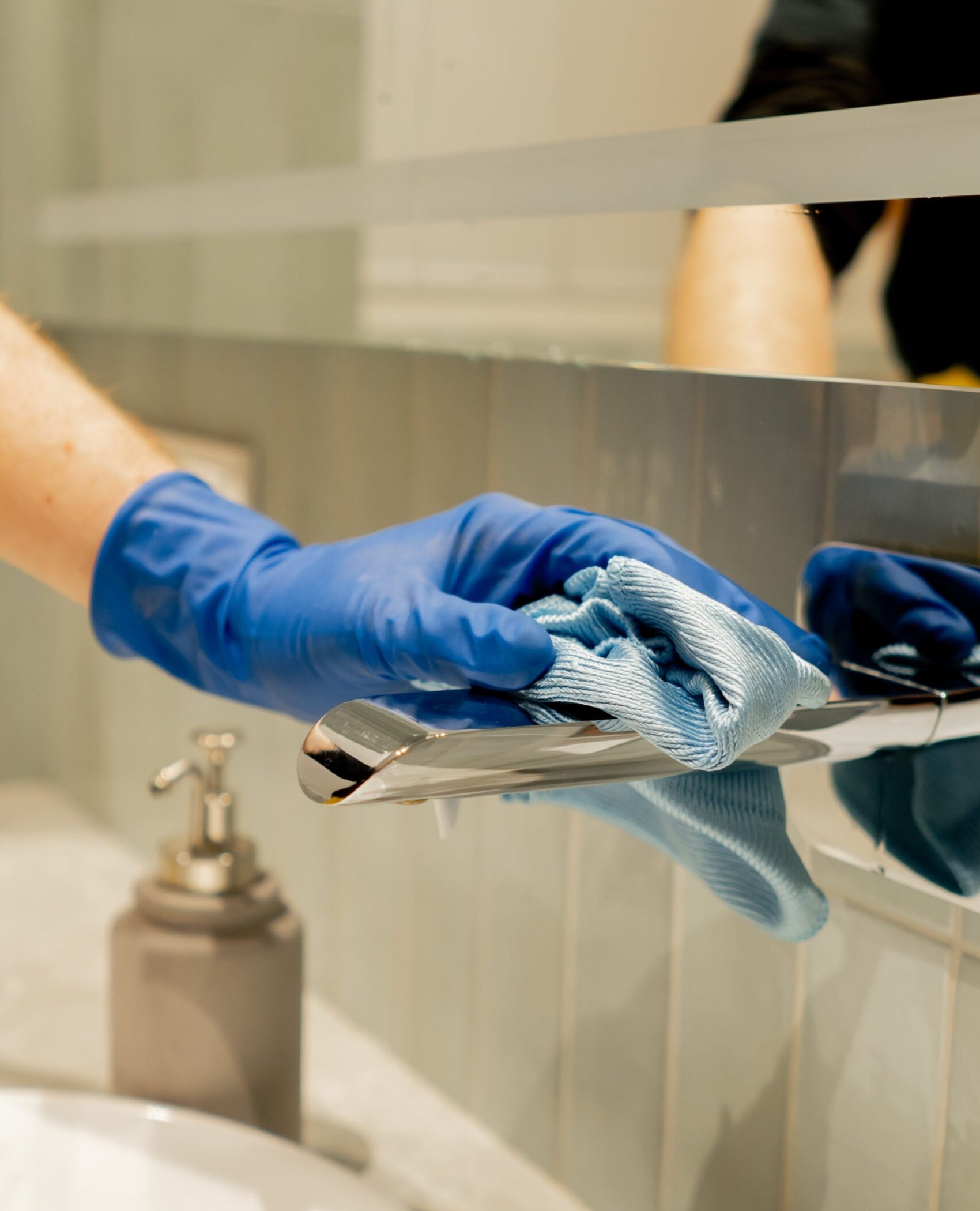 Eine Hand in einem blauen Handschuh reinigt einen Chromhahn mit einem Tuch; Badezimmerreinigung in Hannover