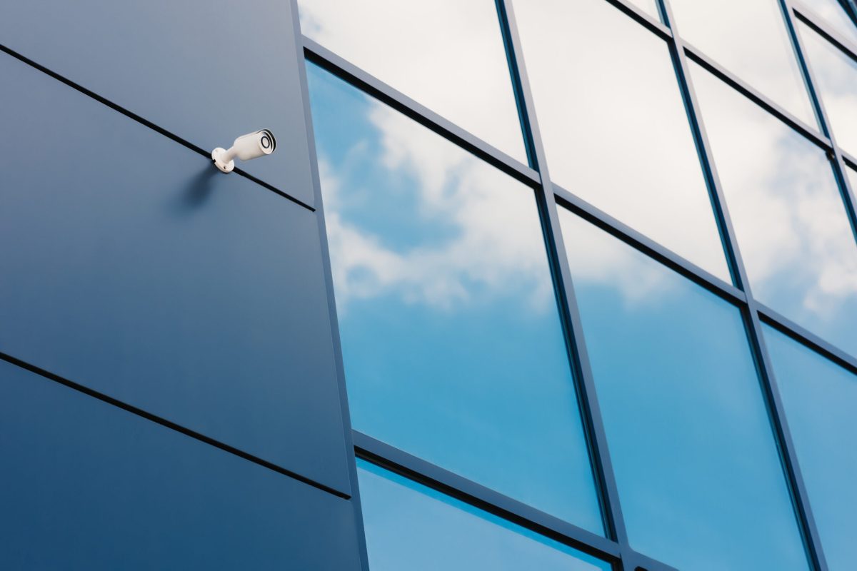 Sicherheitskamera an der Fassade eines modernen Bürogebäudes, dessen blaue Glasfassade den Himmel reflektiert – Ein Service von A&S Service Hannover.